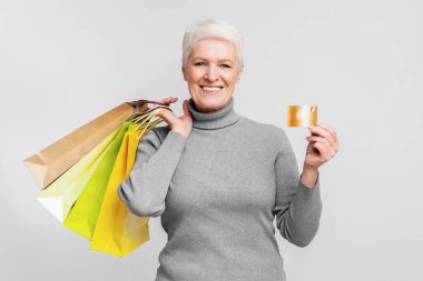 Avrupalı yaşlı bir kadın elinde renkli alışveriş çantaları ve altın bir kredi kartı tutarak gülümsüyor, bu da s3niorlife tüketiciliğini simgeliyor.