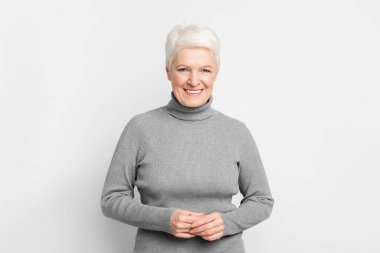 Elleri sımsıkı bağlı neşeli yaşlı Avrupalı kadın, gri arka planda s3niorlife memnuniyetini gösteriyor