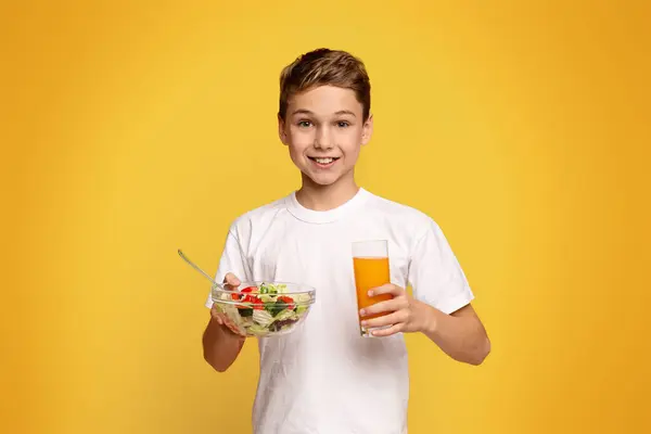 野菜サラダと新鮮なオレンジジュースを食べる笑顔の小さな男の子 オレンジスタジオの背景 — ストック写真
