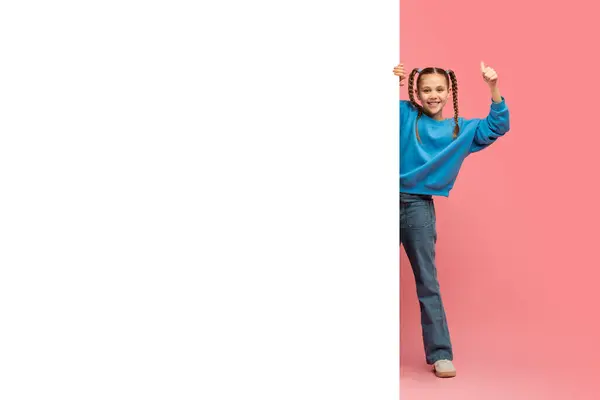 青いセーターとジーンズの陽気な若い女の子は ピンクの背景に対して白いボードの後ろに隠れて 十分なコピースペース ストック写真