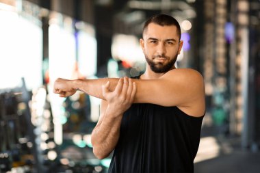 Bir adam ön planda durur, bir kolunu vücuduna uzatır, diğer eliyle dirseğini kavrarken tipik bir kolunu uzatır.