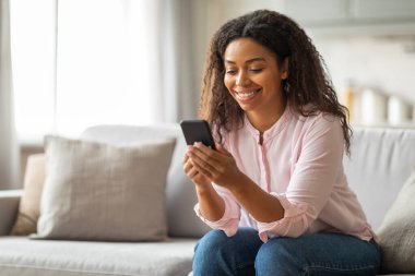 Evde rahat ve rahat bir şekilde, genç siyah bir kadın koltukta oturuyor, telefonundaki mesajları okurken gülümsüyor, rahat, günlük bir anı yansıtıyor.