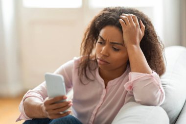 Modern bir oturma odasında, genç siyah bir kadın kanepede oturuyor, akıllı telefonuyla iletişim kurarken yüzündeki endişe ifadesi evde stresli bir gün geçirdiğini gösteriyor..