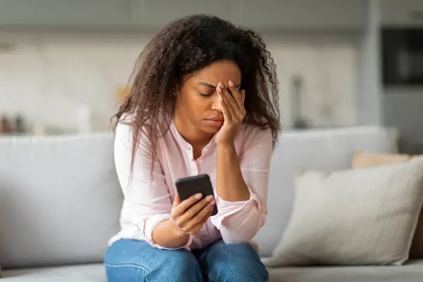 stock image A poignant moment with an african american lady sitting at home, appearing worried as she checks her phone, evoking empathy