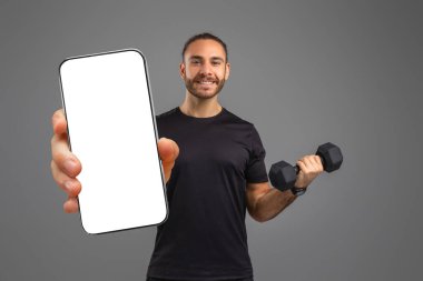 Bir adam elinde cep telefonu ve dambıl tutuyor ve ekranı boş. Telefona dikkatle bakıyor gibi görünüyor. Model, kopyalama alanı.
