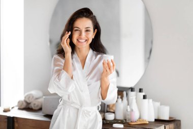 Beyaz bornozlu neşeli bir kadın, krem sürerek, banyoda bin yıllık güzellik ayinlerini sergiliyor.