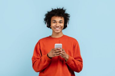 Sıradan giyinen, gülümseyen ve akıllı telefonuna bakan, bağlantıyı ve boş zamanı sembolize eden, mavi üzerine izole edilmiş Afro-Amerikan bir zoomer.