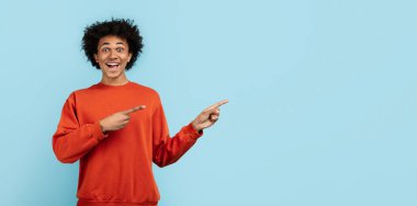 Hevesli, Afro-Amerikalı, kırmızı kazaklı genç bir adam neşeli bir şekilde mavi bir arka plana işaret ediyor.