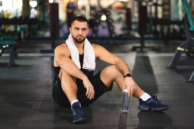 Spor kıyafetleri içindeki bir adam spor salonunun zemininde oturuyor. Etrafı egzersiz ekipmanlarıyla çevrili. Antrenmandan sonra dinleniyor ya da esniyor gibi..