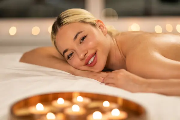 Smiling stunning woman is lying gracefully on top of a bed, placed next to flickering candles. The soft glow of the candles illuminates her features as she relaxes in this cozy setting.