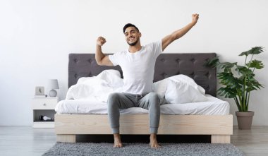 Handsome Happy Young Arab Guy Waking Up In The Morning, Sitting On Bed And Stretching After Good Sleep, Smiling Millennial Middle Eastern Man Having Good Mood, Enjoying Start Of New Day, Copy Space