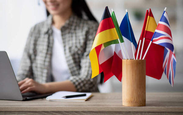 Обрезанный азиатской молодой женщиной учитель иностранного языка, сидящей за рабочим столом и использующей ноутбук, избирательный акцент на международных флагах. Иностранный язык онлайн курс, класс, концепция электронного образования