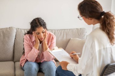 Genç bir kadın, gündüz danışma seansı sırasında rahat bir kanepede otururken, başını ellerinin arasında tutarak sorunlu görünüyor.