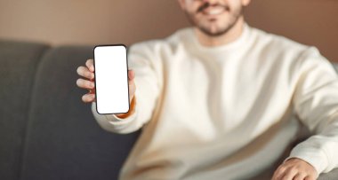 Evdeki koltukta oturan bir adam elinde beyaz ekranlı bir cep telefonu tutuyor, panorama.