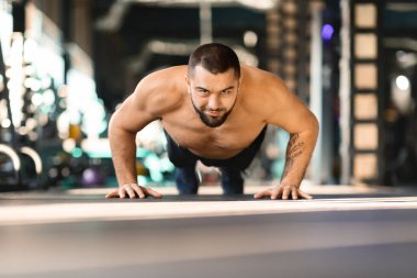 Gömleksiz bir adam spor salonunda şınav çekiyor. Vücudunu indirip kaldırırken kasları kasılmış. Spor aletleri ve arka planda çalışan diğer insanlar aktif atmosfere katkı sağlıyor..