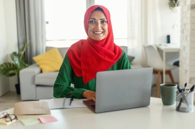 Müslüman kadın bir masada oturuyor, dizüstü bilgisayarına dalmış. Ekran, yazı yazarken, işine odaklanırken ya da elindeki işe odaklanırken yüzünün üzerine yumuşak bir ışık saçıyor..
