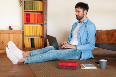 Sıradan giyimli genç bir adam, arka planda renkli kitaplıklar bulunan tahta bir zeminde bağdaş kurarak otururken dizüstü bilgisayarına odaklanıyor. Yanında bir akıllı telefon, bir defter ve bir fincan kahve.