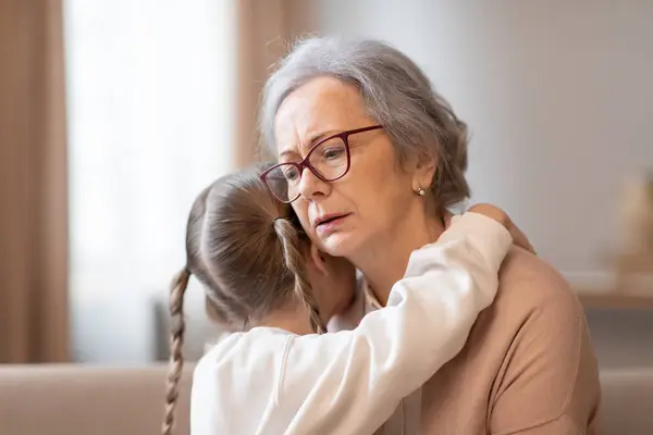 A grandmother with glasses and a caring expression is hugging her young granddaughter, who seems to be seeking comfort or reassurance. They are closely embracing in a warmly lit room