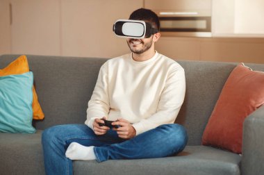 Bir adam kanepeye oturmuş, sanal gerçeklik deneyimine dalmış. Bir VR kulaklık takıyor ve görüntülenen dijital içeriğe odaklanmış gibi görünüyor..