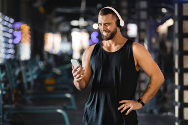Kolsuz bluzlu ve kulaklıklı bir adam spor salonunda akıllı telefonundan şarkı listesi seçerken gülümsüyor. Rahatlamış duruşu ve neşeli ifadesi spor yaparken bir anlık zevk veriyor..