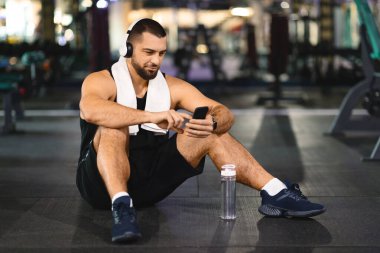 Bir adam cep telefonu ekranına dikkatle bakarken yerde oturuyor. Duruşu cihaza odaklandığını gösteriyor. Çevrede dikkat dağıtıcı bir şey yok..