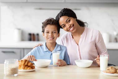 Neşeli bir Afrikalı Amerikalı anne ve oğlu mutfak masasında gülümseyerek bir anlık mutluluk ve beraberlik sergilerler.