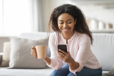 Kahve fincanı kullanan neşeli Afro-Amerikalı kadın, rahatlama ve ev hayatı örneğini veriyor.