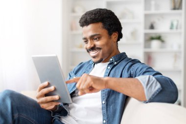 Gülümseyen Afro-Amerikalı adam evde dijital tablet kullanıyor, boş zamanlarını ya da internet gezilerini öneriyor.