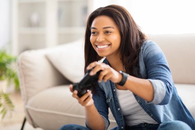 Afrikalı Amerikalı bir kadın kanepeye oturmuş elinde uzaktan kumandayla video oyunları oynuyor ve gülümsüyor.