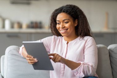 An approachable black woman holds a tablet, her smile suggesting enjoyment or satisfaction from the content shes viewing at home clipart