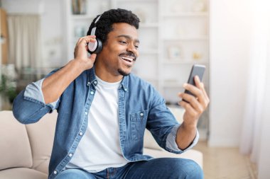 Afro-Amerikalı, kulaklıklı siyahi bir adam evinde neşeyle dans ediyor, akıllı telefonuyla müzik dinliyor, mutlu hissediyor.