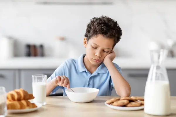 In this image, a bored African American boy sits at the kitchen table, uninterested in his breakfast, highlighting a common family morning scenario