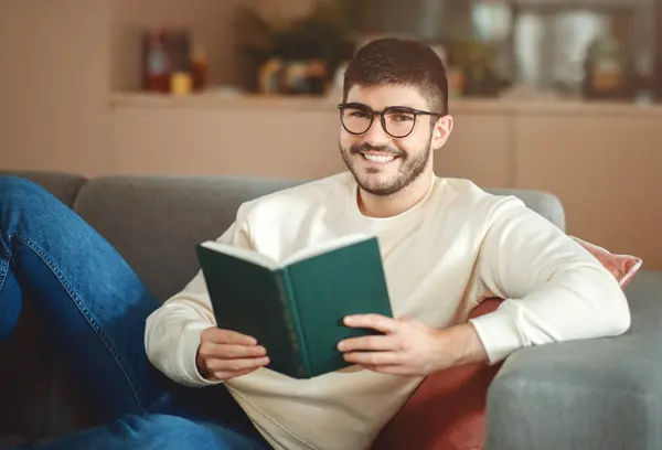 眼鏡をかぶった男性がソファに座り 手元にある本に囲まれています 部屋は彼が読書に吸収されたページを飛び越えて落ち着いているように見えます — ストック写真