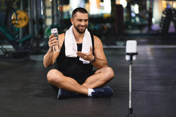 A cheerful man sits cross-legged on the gym floor, holding a water bottle and gesturing towards a smartphone on a tripod, seemingly engaging with his audience during a break