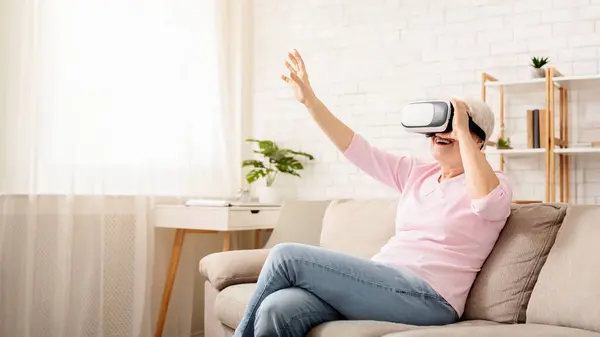 Stock image Excited senior woman enjoying cyber world, using VR glasses at home, free space