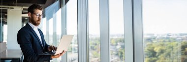 Başarılı orta yaşlı iş adamı, elinde dizüstü bilgisayarla pencerenin yanındaki modern ofis binasında duruyor, kopya alanı olan bir panorama. Bilgisayarda çalışan resmi takım elbiseli ciddi girişimci.