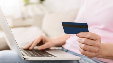 Masada oturan bir kadın bir elinde kredi kartı, diğer elinde dizüstü bilgisayar kullanıyor. Muhtemelen online alışveriş veya ödeme yapıyor..