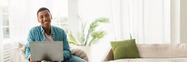 Содержание афроамериканец улыбается с ноутбуком на диване, подходит для веб-баннера и копирования пространства