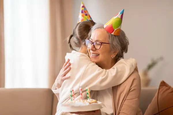 stock image A tender moment captured as an older woman embraces a young girl, both wearing celebratory birthday hats. The affection and joy between them are evident in the heartfelt hug shared.