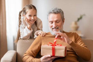 Sahnede, yaşlı bir adam ve genç bir kız birlikte duruyorlar, paketlenmiş bir hediye tutuyorlar. İkisi de gülümsüyor ve hediyelerini verirken bir anlık sevinçlerini paylaşıyor gibi görünüyorlar..
