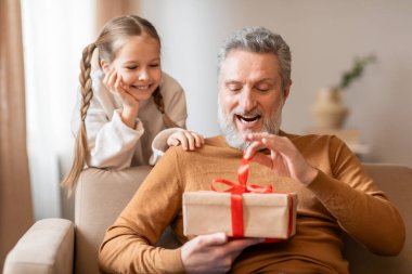 Yaşlı bir adam ve genç bir kız bir kanepede oturuyorlar, ikisi de aralarında paketlenmiş hediyeye heyecanlı bir şekilde bakıyorlar. Büyükbaba gülümsüyor ve küçük kız meraklı görünüyor. Oda rahat ve aydınlık..