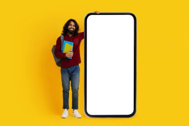 Sakal öğrencisi olan neşeli Hintli adam elinde renkli kitaplarla sol tarafta duruyor ve büyük, boş bir ekran akıllı telefonuna yaslanıyor.