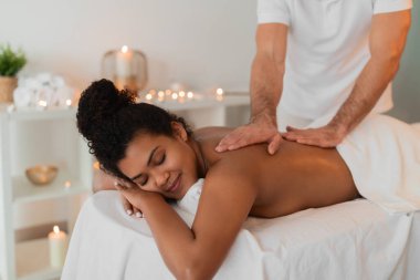 Profesyonel sırt masajının tadını çıkaran Afro-Amerikan bir bayan, yumuşak ışıklandırmalı ve rahatlatıcı bir atmosferi olan bir kaplıcada uzanıyor.