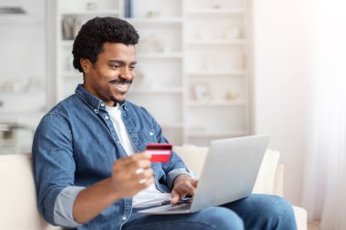 Neşeli, Afro-Amerikalı bir adam bilgisayarını ve kredi kartını kullanarak rahatça evden alışveriş yapıyor ve online alışveriş yapıyor.