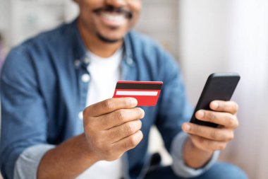 Evinden online alışveriş yapan, kredi kartını ve cep telefonunu gösteren, modern tüketiciliğin bir temsilcisi olan Afrikalı bir Amerikalının yakın çekimi,