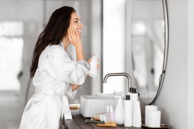 Beyaz cüppeli bir kadın iyi aydınlatılmış bir banyoda ayna ve modern lavabo ile cilt ürününü uygular.