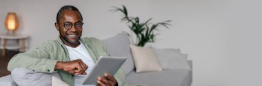Gözlüklü gülümseyen Afro-Amerikan bir adam ve tablet üzerinde günlük videolar izleyip oturma odasının içindeki sosyal ağlarda sohbet ediyor. Yeni uygulama, uzaktan kumandayı evde çalıştır, blog, kopyalama alanı olan panorama