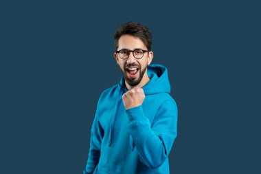 Canlı, gözlüklü genç bir adam, parlak mavi kapüşonlu bir kapüşonlu ile orta sahada yakalandı. Yumruğunu sıkar ve ağzını sonuna kadar açar. Heyecan ve destek jesti olarak.