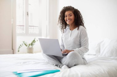 İspanyol bir kadın, dizüstü bilgisayar kullanmaya odaklanmış bir şekilde yatakta oturuyor. Ekranda veri göstererek klavyeye yazar. Oda iyi aydınlatılmış, basit ve temiz bir dekorla..