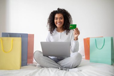 Neşeli, genç bir İspanyol kadın dizüstü bilgisayarıyla yatağında oturuyor, etrafında alışveriş torbaları olan bir kredi kartı gösteriyor, başarılı bir online alışveriş oturumu öneriyor..